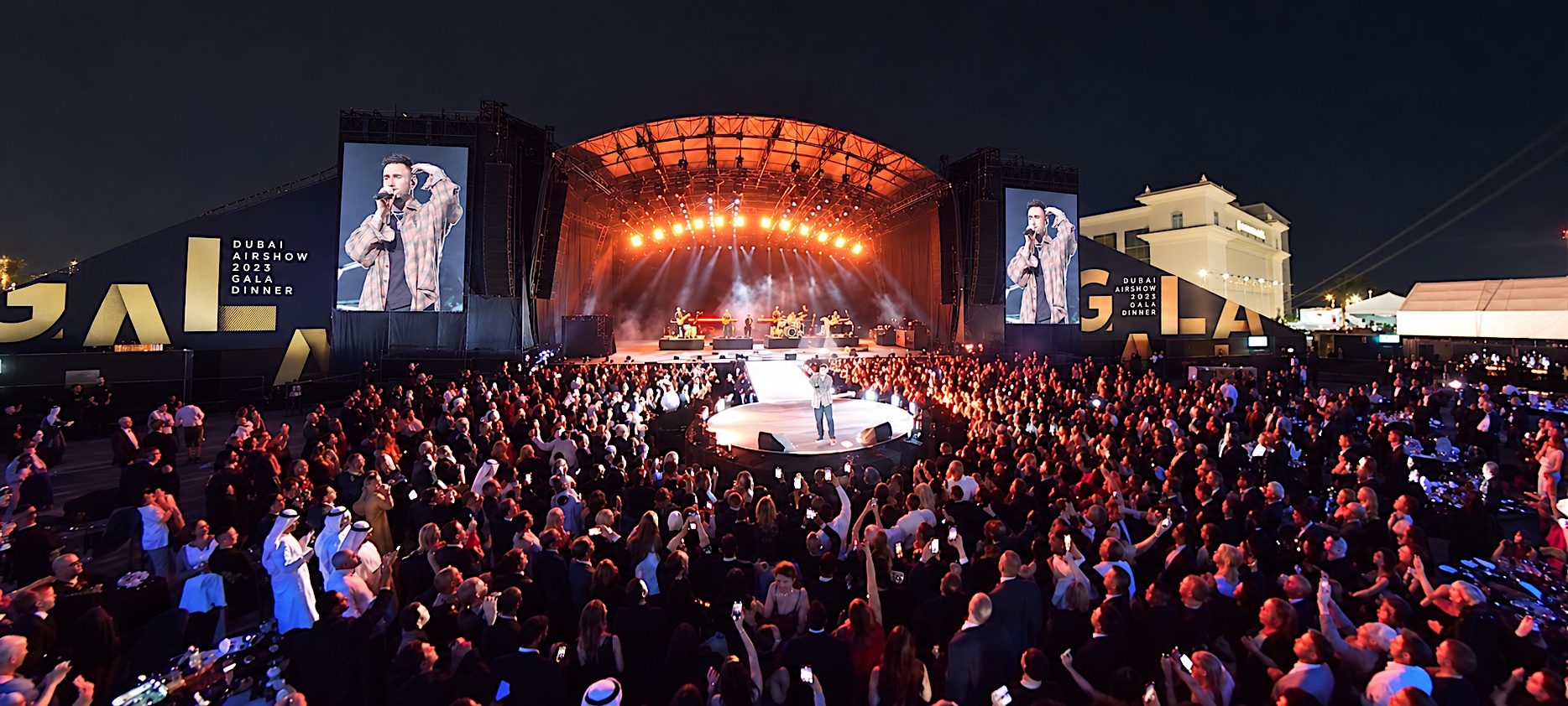 A singer on a round stage at a gala dinner, his close-up on the big screens either side, the crowd filming on their phones