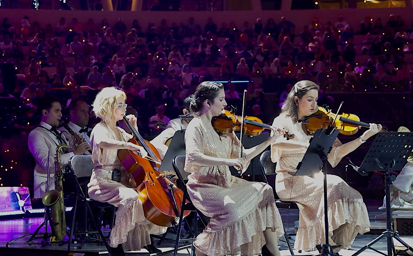 A string section in gold dresses playing on stage in front of a packed arena audience