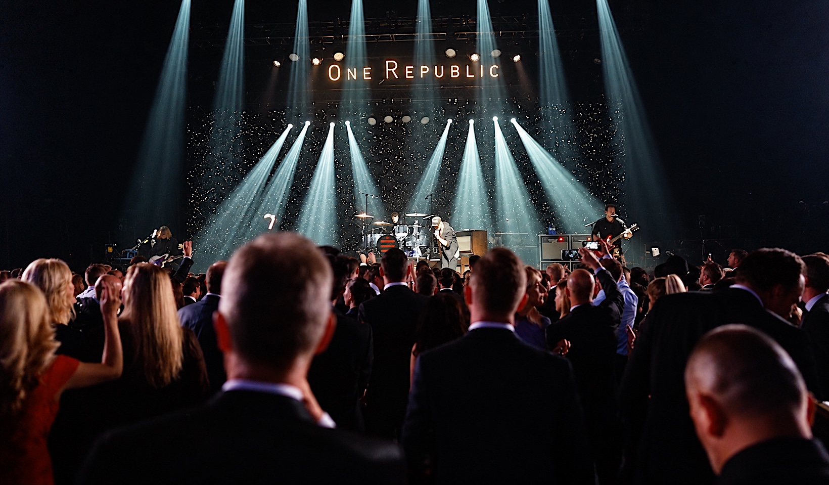 OneRepublic on stage under beams of concert light, a gala crowd in the foreground
