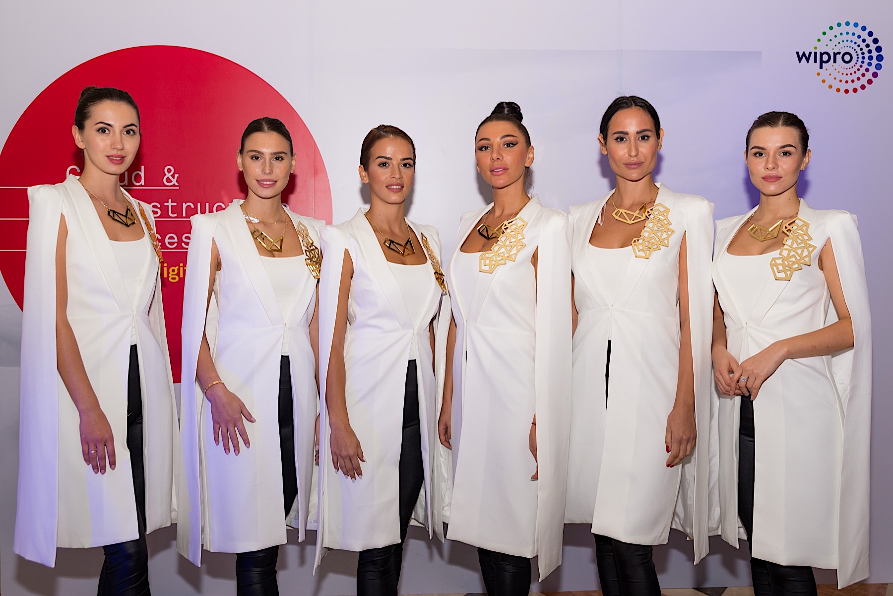 Six models in white cape coats and gold geometric necklaces at a corporate event